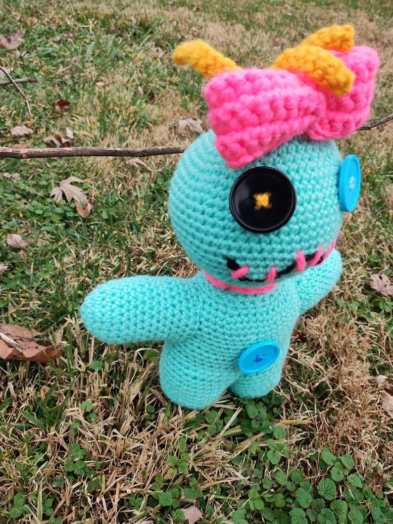 Op de afbeelding: Een gehaakte pop in turquoise met een roze strik, gele antennes en knoopogen. De pop heeft een gestikte roze mond en blauwe knoopaccenten. Het wordt buiten op gras gepresenteerd.