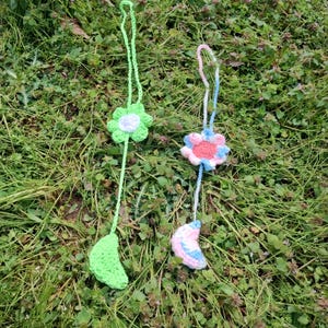May include: Two handmade crochet pendants. One features a green flower with a white center and a green leaf. The other has a pink, blue, and white flower with a crescent moon charm. Both are suspended by a green and pink cord, respectively.