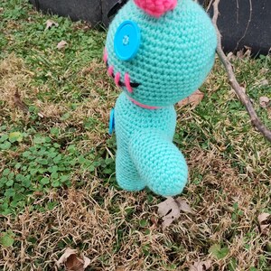 Op de afbeelding: Een gehaakte pop in turquoise met een roze hoofdband en accenten. De pop heeft knoopogen en roze stikseldetails. Hij staat op gras en bladeren. De pop is ongeveer 15 cm hoog.
