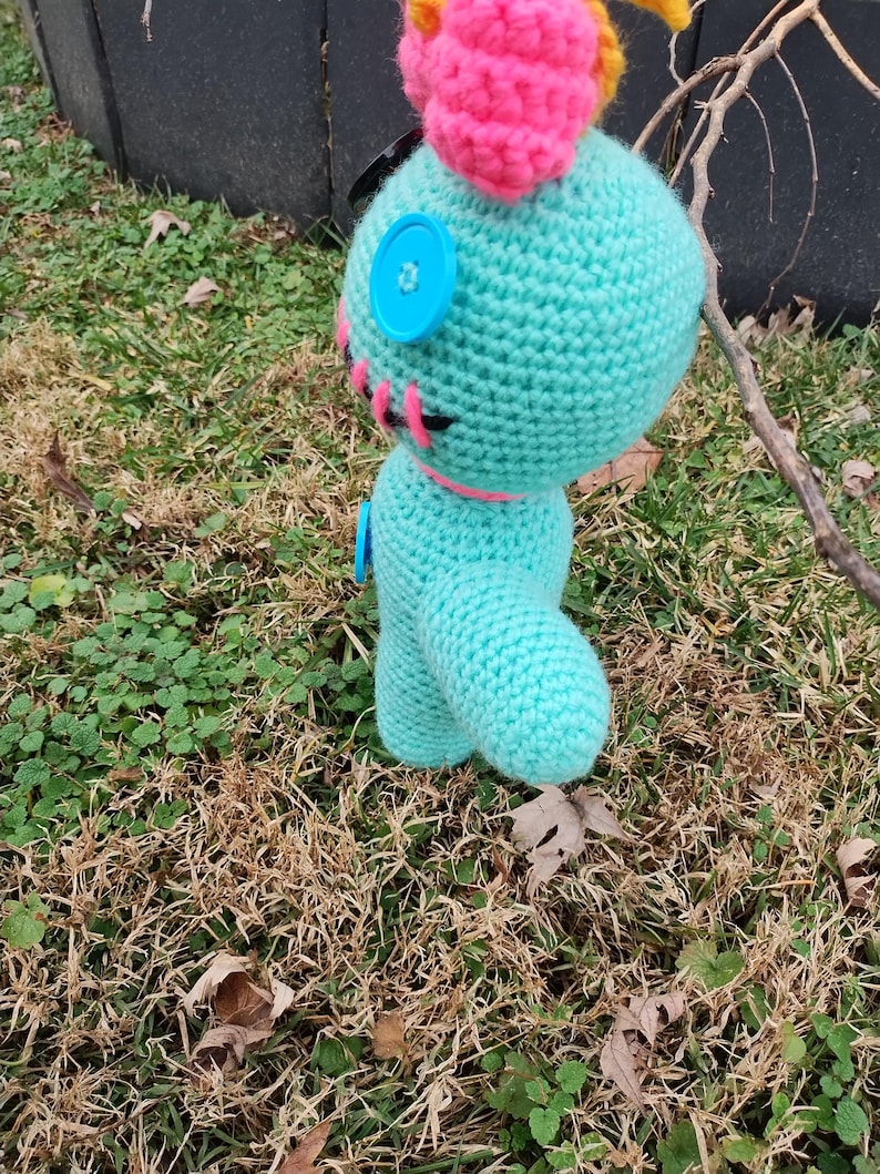 Op de afbeelding: Een gehaakte pop in turquoise met een roze hoofdband en accenten. De pop heeft knoopogen en roze stikseldetails. Hij staat op gras en bladeren. De pop is ongeveer 15 cm hoog.