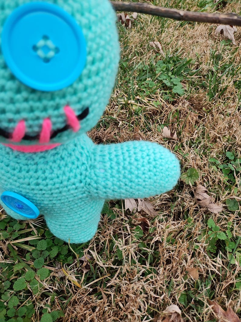 Op de afbeelding: Een turquoise gehaakte pop met grote blauwe knoopogen en een roze gestikte mond. De pop heeft een gestikt ontwerp op het gezicht en een kleine uitgestrekte arm. De pop staat tegen een achtergrond van droog gras en bladeren.