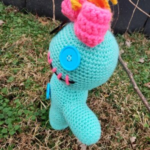 Op de afbeelding: Een handgemaakte turquoise gehaakte pop met een roze strik en gele accenten. De pop heeft blauwe knoopogen en roze gestikte details. De pop staat op een bed van gras.