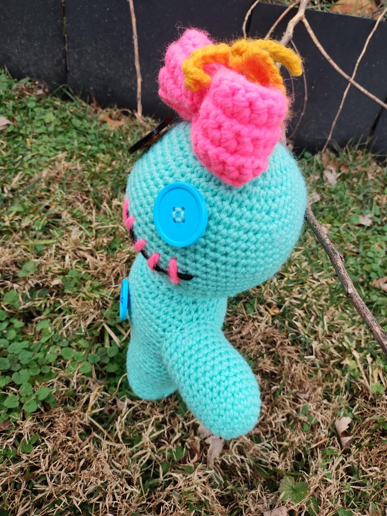 Op de afbeelding: Een handgemaakte turquoise gehaakte pop met een roze strik en gele accenten. De pop heeft blauwe knoopogen en roze gestikte details. De pop staat op een bed van gras.