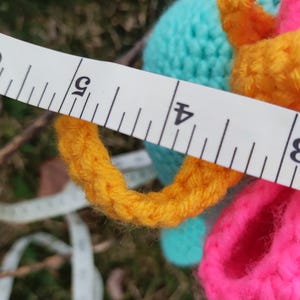 Op de afbeelding: Een close-up van een kleurrijk gehaakt item met een meetlint. Het item heeft heldere kleuren, waaronder geel, roze en turquoise. Het meetlint toont een meting van ongeveer 10 cm.