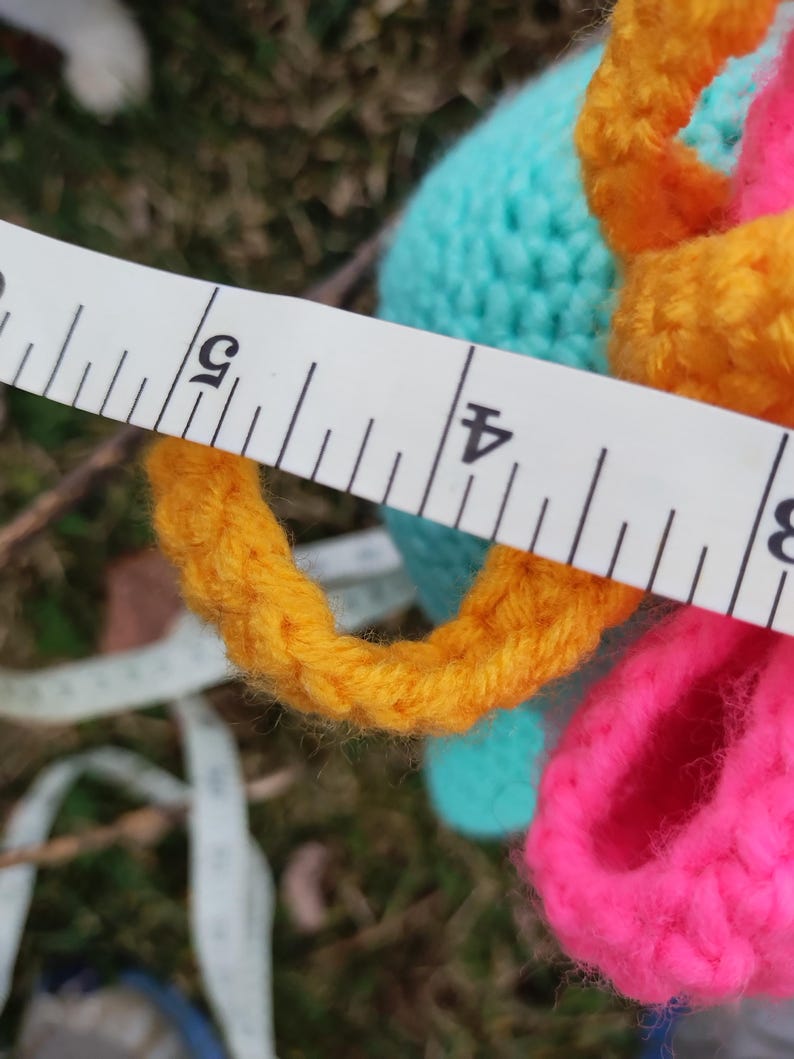Op de afbeelding: Een close-up van een kleurrijk gehaakt item met een meetlint. Het item heeft heldere kleuren, waaronder geel, roze en turquoise. Het meetlint toont een meting van ongeveer 10 cm.