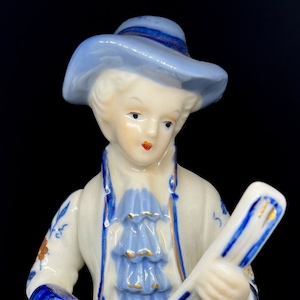 May include: A porcelain figurine of a person wearing a blue hat and coat, playing a stringed instrument. The figurine is painted in blue and white with gold accents.