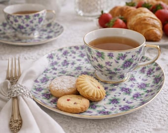 Può includere: Una tazza e un piattino con motivo floreale viola, pieni di tè, insieme a un piatto con biscotti. Un tovagliolo bianco con una forchetta decorativa e un bouquet di rose rosa creano una classica scena del tè del pomeriggio.