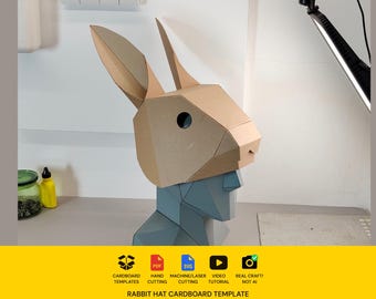 Könnte beinhalten: Eine geometrische Hasenkopfmaske aus beigefarbenem Karton mit hohen, spitzen Ohren und kreisförmigen Augenausschnitten. Die Maske wird auf einer blauen, geometrischen Büste präsentiert. Unter dem Bild steht der Text: Cardboard Templates, Hand Cutting, Machine/Laser Cutting, Video Tutorial, Real Craft! Not AI, Rabbit Hat Cardboard Template.