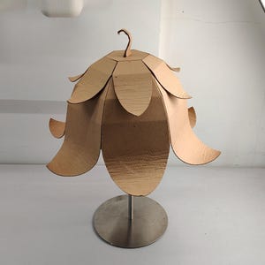 May include: A light brown cardboard lamp shade shaped like a flower with multiple overlapping petals. The shade is mounted on a thin, vertical silver metal rod attached to a circular, flat silver metal base, creating a structured, layered appearance.