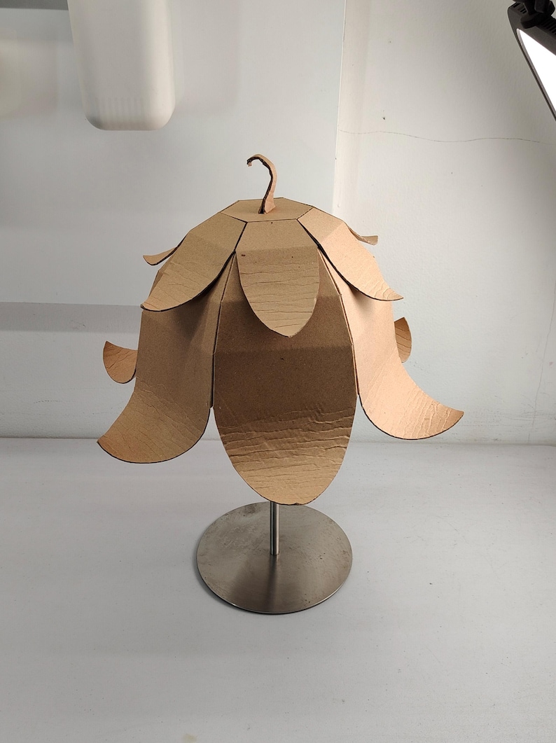 May include: A light brown cardboard lamp shade shaped like a flower with multiple overlapping petals. The shade is mounted on a thin, vertical silver metal rod attached to a circular, flat silver metal base, creating a structured, layered appearance.