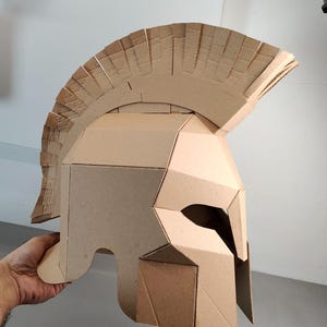 May include: A tan cardboard helmet featuring a prominent crest along the top, angular side panels, and a narrow eye opening. The construction consists of multiple layered pieces of corrugated cardboard joined together to form a rigid structure.