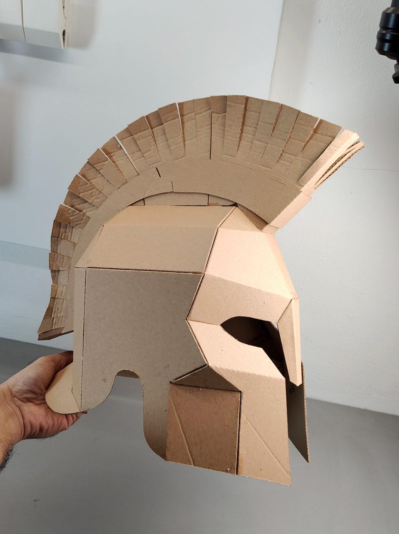 May include: A tan cardboard helmet featuring a prominent crest along the top, angular side panels, and a narrow eye opening. The construction consists of multiple layered pieces of corrugated cardboard joined together to form a rigid structure.