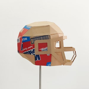 以下が含まれることがあります： シリアルボックスで作られた段ボール製のフットボールヘルメット。ヘルメットは茶色で、赤い部分に「Instant Spud」と「ME!」の文字が書かれた青と赤のデザインが施されています。