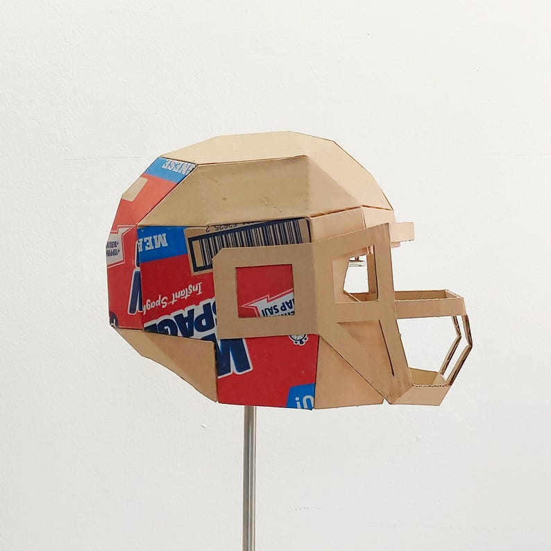 以下が含まれることがあります： シリアルボックスで作られた段ボール製のフットボールヘルメット。ヘルメットは茶色で、赤い部分に「Instant Spud」と「ME!」の文字が書かれた青と赤のデザインが施されています。