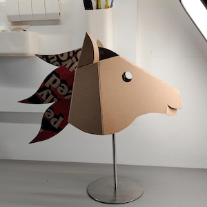Puede incluir: Una escultura de cabeza de caballo hecha de cart&oacute;n marr&oacute;n, montada sobre un soporte de metal plateado. La crin presenta segmentos superpuestos de papel impreso en rojo, negro y blanco con texto visible. La cabeza tiene un recorte circular para el ojo.