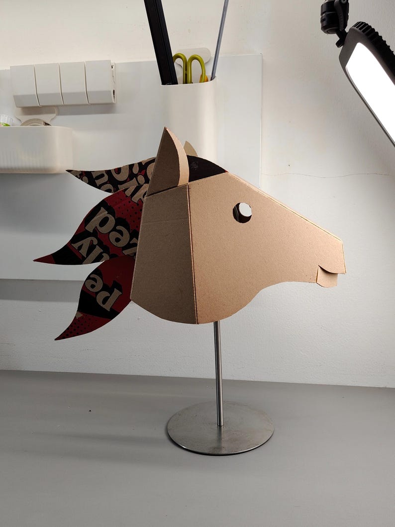 Puede incluir: Una escultura de cabeza de caballo hecha de cart&oacute;n marr&oacute;n, montada sobre un soporte de metal plateado. La crin presenta segmentos superpuestos de papel impreso en rojo, negro y blanco con texto visible. La cabeza tiene un recorte circular para el ojo.