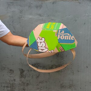 K&ouml;nnte beinhalten: Ein Papphelm aus recycelten Lebensmittelverpackungen. Der Helm ist gr&uuml;n, blau und braun mit dem Text "Makaron Cepat Saji" und "La Fonte" darauf.