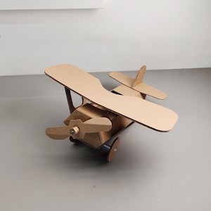 May include: A biplane model constructed from brown corrugated cardboard. The aircraft features a main fuselage, two stacked wings, a front-mounted propeller, a vertical tail fin, a horizontal stabilizer, and two circular wheels attached to the base.