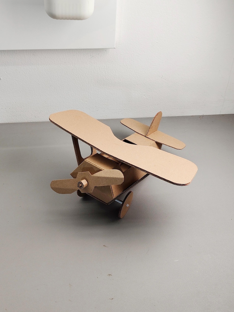 May include: A biplane model constructed from brown corrugated cardboard. The aircraft features a main fuselage, two stacked wings, a front-mounted propeller, a vertical tail fin, a horizontal stabilizer, and two circular wheels attached to the base.