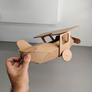 May include: A handcrafted model aeroplane constructed from brown corrugated cardboard. The structure features a main fuselage, a single top wing supported by struts, a tail section, and two circular wheels attached to the sides of the body.
