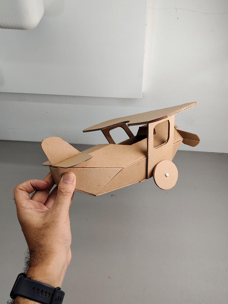 May include: A handcrafted model aeroplane constructed from brown corrugated cardboard. The structure features a main fuselage, a single top wing supported by struts, a tail section, and two circular wheels attached to the sides of the body.