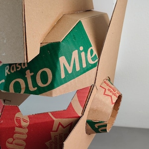 May include: A geometric sculpture constructed from recycled brown cardboard packaging. The structure features angular panels with green and red printed sections, displaying text including 'Rasa', 'oto Mie', and 'Lengkap Dengan Saus Cabe'.