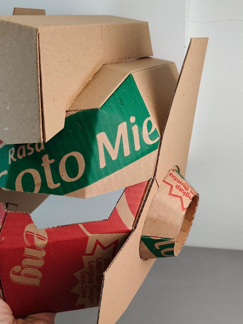 May include: A geometric sculpture constructed from recycled brown cardboard packaging. The structure features angular panels with green and red printed sections, displaying text including 'Rasa', 'oto Mie', and 'Lengkap Dengan Saus Cabe'.