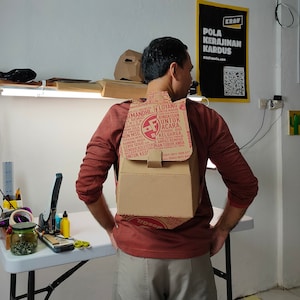 May include: A person wearing a red shirt with a cardboard box strapped to their back. The box is decorated with red and white text in Indonesian. The text reads "Sastibis Imandhi Basmat Non MSG. Kambing Muda Lebih Sehat Rendam Untuk Kes Loyang Bingkisan Untuk Acara Kerabad Atan Keluarga Ampas Rempah Anda (18Y) 0811 1699 817 (800) 0811 85".