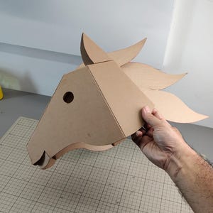 Puede incluir: Una m&aacute;scara de cabeza de caballo de cart&oacute;n marr&oacute;n claro con un dise&ntilde;o geom&eacute;trico y angular, que presenta una oreja puntiaguda, un recorte circular para el ojo y una crin fluida compuesta por varios segmentos de cart&oacute;n curvos.