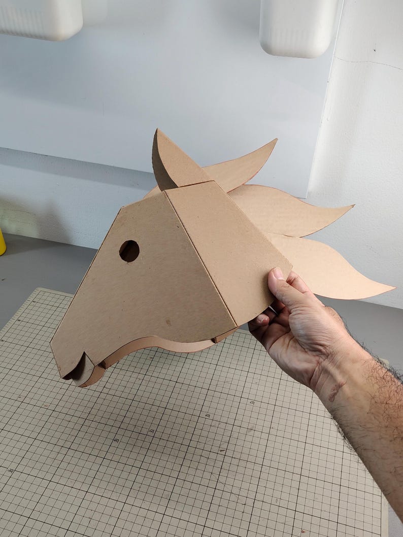 Puede incluir: Una m&aacute;scara de cabeza de caballo de cart&oacute;n marr&oacute;n claro con un dise&ntilde;o geom&eacute;trico y angular, que presenta una oreja puntiaguda, un recorte circular para el ojo y una crin fluida compuesta por varios segmentos de cart&oacute;n curvos.