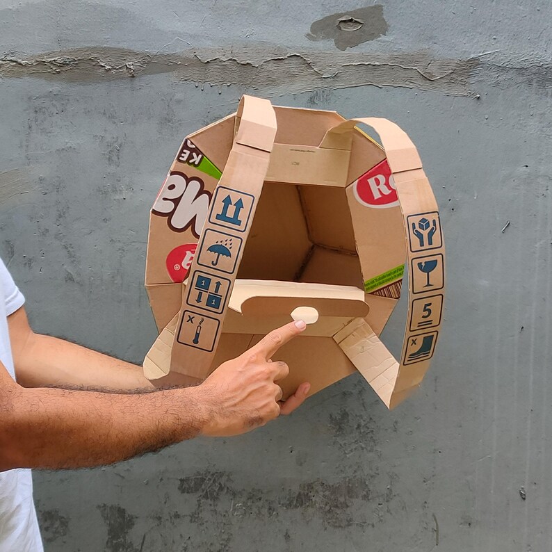 K&ouml;nnte beinhalten: Ein Rucksack aus Pappe, hergestellt aus recycelten Verpackungen. Der Rucksack hat zwei Tr&auml;ger und eine Klappe mit einer kleinen &Ouml;ffnung. Die Verpackung enth&auml;lt verschiedene Symbole und Texte, darunter "KE" und "RO".