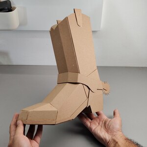 Puede incluir: Una escultura tridimensional de bota vaquera construida con cart&oacute;n color canela. La bota presenta un dise&ntilde;o geom&eacute;trico y facetado con una espuela prominente unida al &aacute;rea del tal&oacute;n. La estructura est&aacute; ensamblada con paneles planos y costuras.