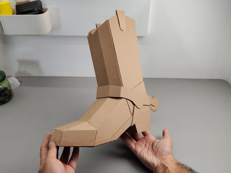 Puede incluir: Una escultura tridimensional de bota vaquera construida con cart&oacute;n color canela. La bota presenta un dise&ntilde;o geom&eacute;trico y facetado con una espuela prominente unida al &aacute;rea del tal&oacute;n. La estructura est&aacute; ensamblada con paneles planos y costuras.