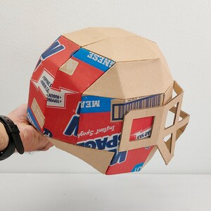 以下が含まれることがあります： インスタントスパゲッティの箱から作られた段ボール製のフットボールヘルメット。ヘルメットは茶色で、赤、青、白の箱が取り付けられています。箱には「インスタントスパゲッティ」と「食事」の文字が印刷されています。