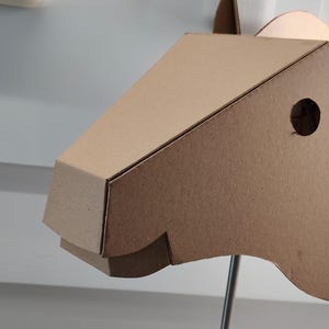 Puede incluir: Una escultura de cabeza de caballo en tres dimensiones construida con cart&oacute;n de color marr&oacute;n claro. El dise&ntilde;o geom&eacute;trico presenta un hocico puntiagudo, un recorte circular para el ojo y una oreja erguida. La estructura est&aacute; montada sobre una varilla de metal.