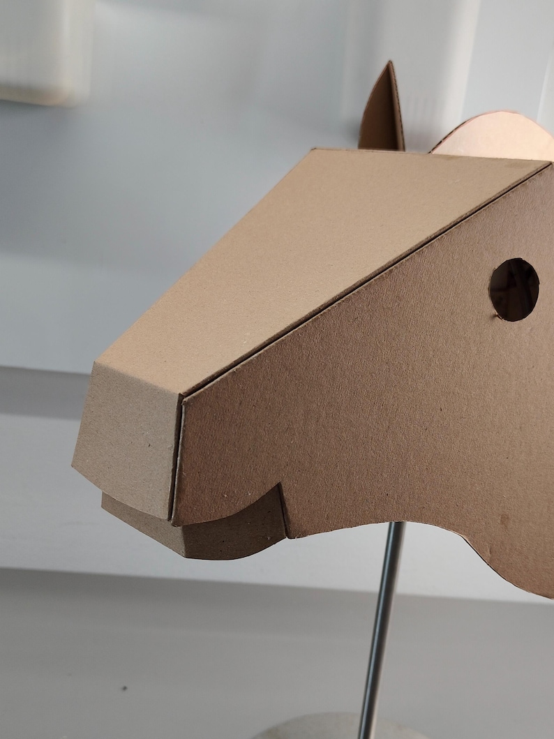 Puede incluir: Una escultura de cabeza de caballo en tres dimensiones construida con cart&oacute;n de color marr&oacute;n claro. El dise&ntilde;o geom&eacute;trico presenta un hocico puntiagudo, un recorte circular para el ojo y una oreja erguida. La estructura est&aacute; montada sobre una varilla de metal.