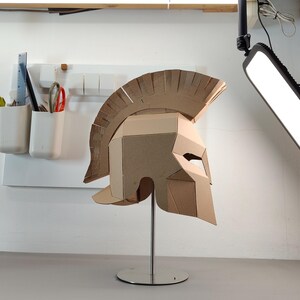 May include: A tan cardboard Spartan-style helmet featuring a prominent crest and angular face guard, mounted on a silver-colored metal stand with a circular base. The helmet is constructed from layered corrugated cardboard panels.