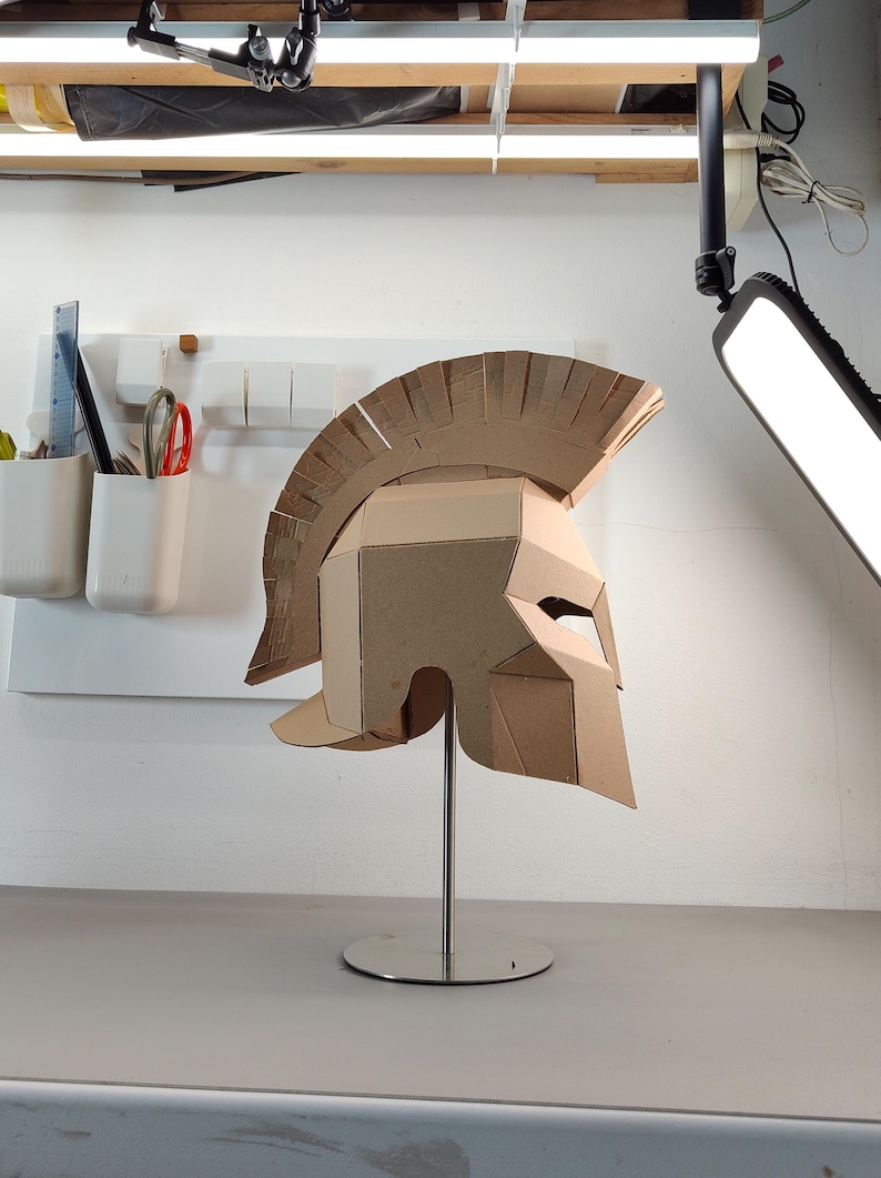 May include: A tan cardboard Spartan-style helmet featuring a prominent crest and angular face guard, mounted on a silver-colored metal stand with a circular base. The helmet is constructed from layered corrugated cardboard panels.