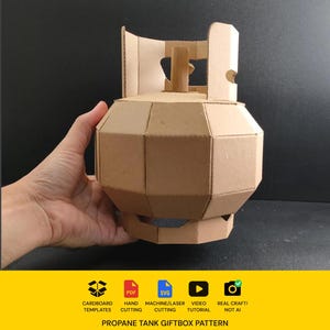 May include: A tan cardboard model of a propane tank with a faceted, geometric body and a protective collar at the top. The base features a circular cutout design. Below the model, text reads: CARDBOARD TEMPLATES, PDF, SVG, VIDEO TUTORIAL, REAL CRAFT! NOT AI, PROPANE TANK GIFTBOX PATTERN.