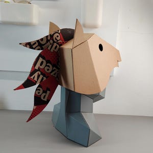 Puede incluir: Una escultura geom&eacute;trica de cabeza de caballo hecha de cart&oacute;n color canela con una melena de papel estampada en rojo oscuro y negro que presenta texto impreso. La secci&oacute;n del cuello y el pecho est&aacute; hecha de paneles geom&eacute;tricos gris claro.