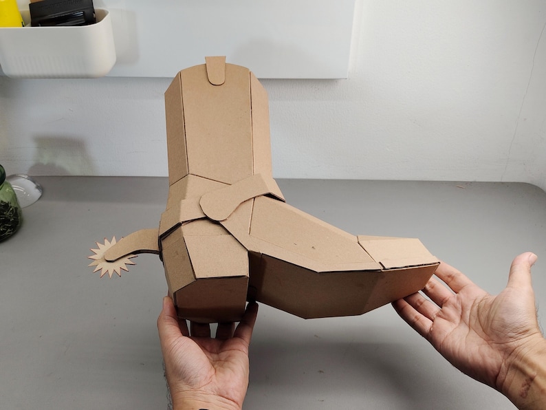 Puede incluir: Una escultura tridimensional de bota vaquera construida con cart&oacute;n marr&oacute;n. La bota presenta un dise&ntilde;o geom&eacute;trico y facetado con una espuela prominente unida al tal&oacute;n, mostrando l&iacute;neas limpias y angulares con una estructura de papel.