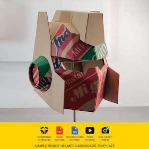May include: A robot helmet constructed from recycled cardboard featuring red, green, and white printed branding text including Mi Instant and Soto. The helmet has a geometric design with a circular side piece and a wide visor opening for the face.