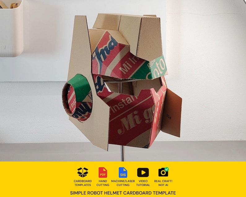 May include: A robot helmet constructed from recycled cardboard featuring red, green, and white printed branding text including Mi Instant and Soto. The helmet has a geometric design with a circular side piece and a wide visor opening for the face.