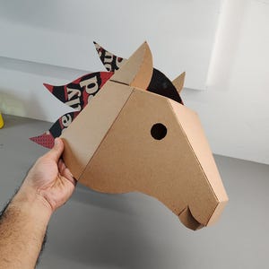 Puede incluir: Una m&aacute;scara de cabeza de caballo en tres dimensiones construida con cart&oacute;n color canela. La crin presenta un patr&oacute;n gr&aacute;fico en negro, rojo y blanco con texto visible. La m&aacute;scara tiene un dise&ntilde;o geom&eacute;trico con un orificio circular.