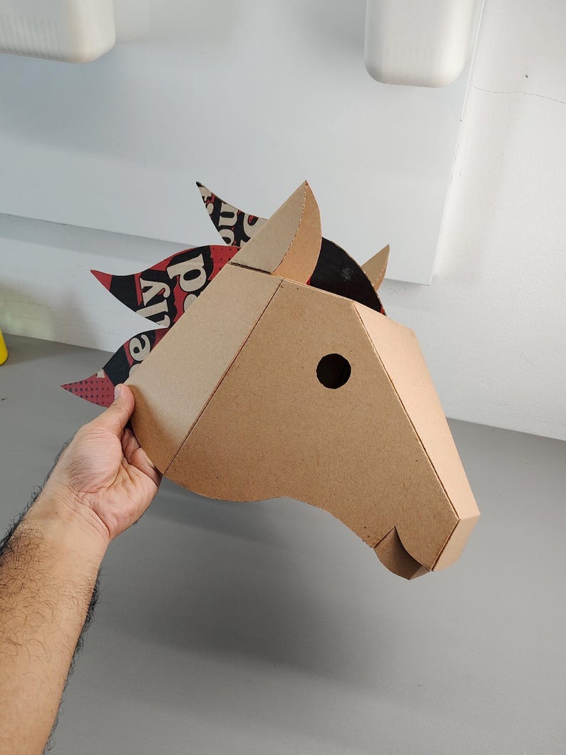 Puede incluir: Una m&aacute;scara de cabeza de caballo en tres dimensiones construida con cart&oacute;n color canela. La crin presenta un patr&oacute;n gr&aacute;fico en negro, rojo y blanco con texto visible. La m&aacute;scara tiene un dise&ntilde;o geom&eacute;trico con un orificio circular.
