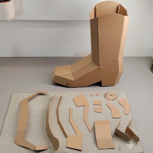 Puede incluir: Una bota de vaquero tridimensional construida con paneles de cart&oacute;n color canela, acompa&ntilde;ada de una colecci&oacute;n de piezas de cart&oacute;n planas y precortadas, incluyendo una espuela en forma de estrella, varias formas geom&eacute;tricas y tiras curvas.