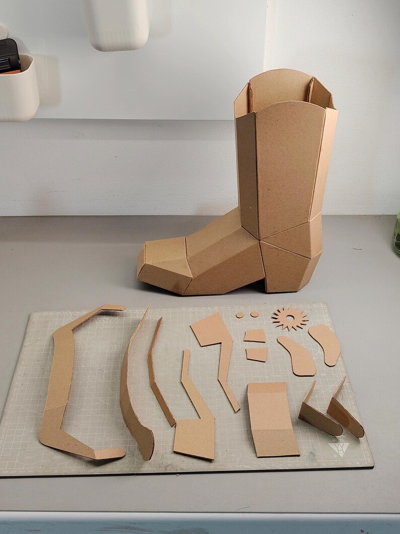 Puede incluir: Una bota de vaquero tridimensional construida con paneles de cart&oacute;n color canela, acompa&ntilde;ada de una colecci&oacute;n de piezas de cart&oacute;n planas y precortadas, incluyendo una espuela en forma de estrella, varias formas geom&eacute;tricas y tiras curvas.