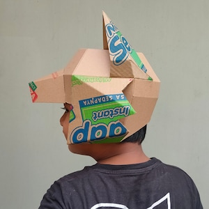 May include: A handmade cardboard helmet with a geometric design. The helmet is constructed from recycled cardboard boxes, featuring printed text and colorful accents. The design includes a pointed top and angular shapes, showcasing a creative, DIY aesthetic.
