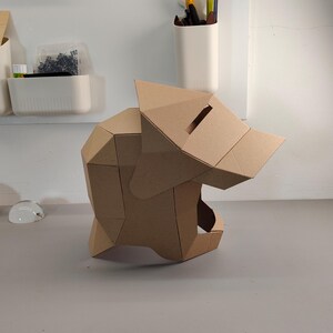 May include: A geometric, cardboard sculpture of a wolf's head. The sculpture is constructed from folded, tan-colored cardboard pieces, with a slot on top. The wolf's head is positioned on a gray surface.