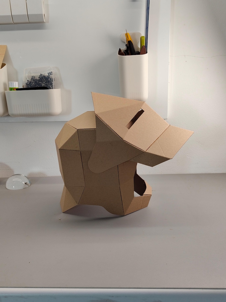 May include: A geometric, cardboard sculpture of a wolf's head. The sculpture is constructed from folded, tan-colored cardboard pieces, with a slot on top. The wolf's head is positioned on a gray surface.